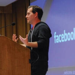 A man speaking at a podium in front of a screen displaying the Facebook logo.